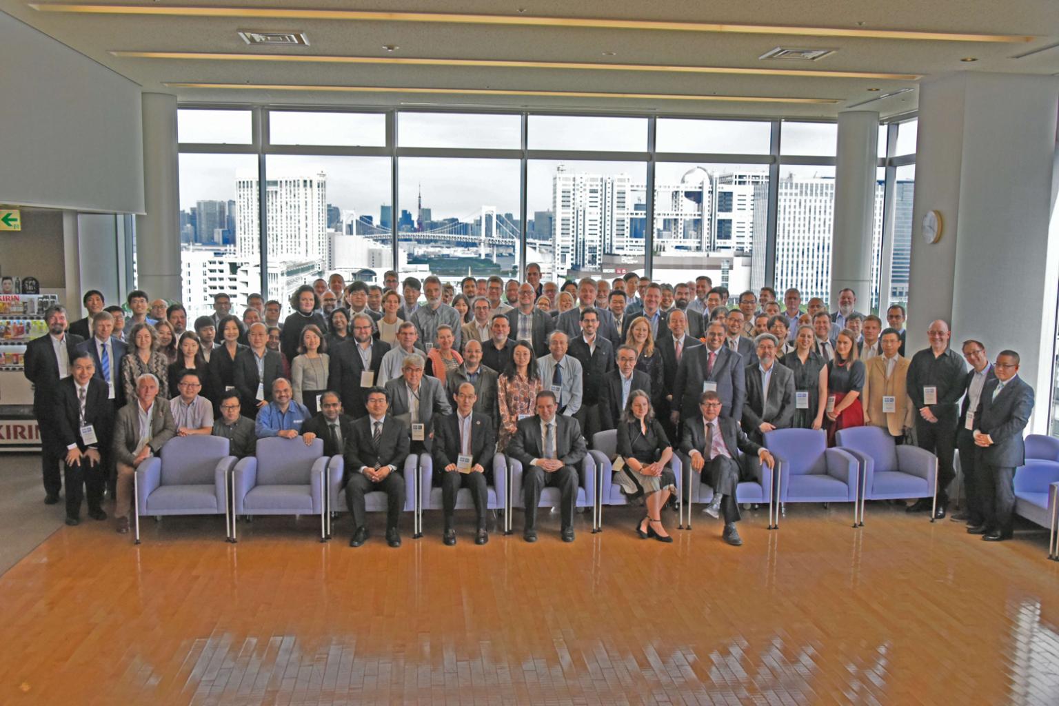 The participants of a large meeting standing in front of a view of Tokyo.